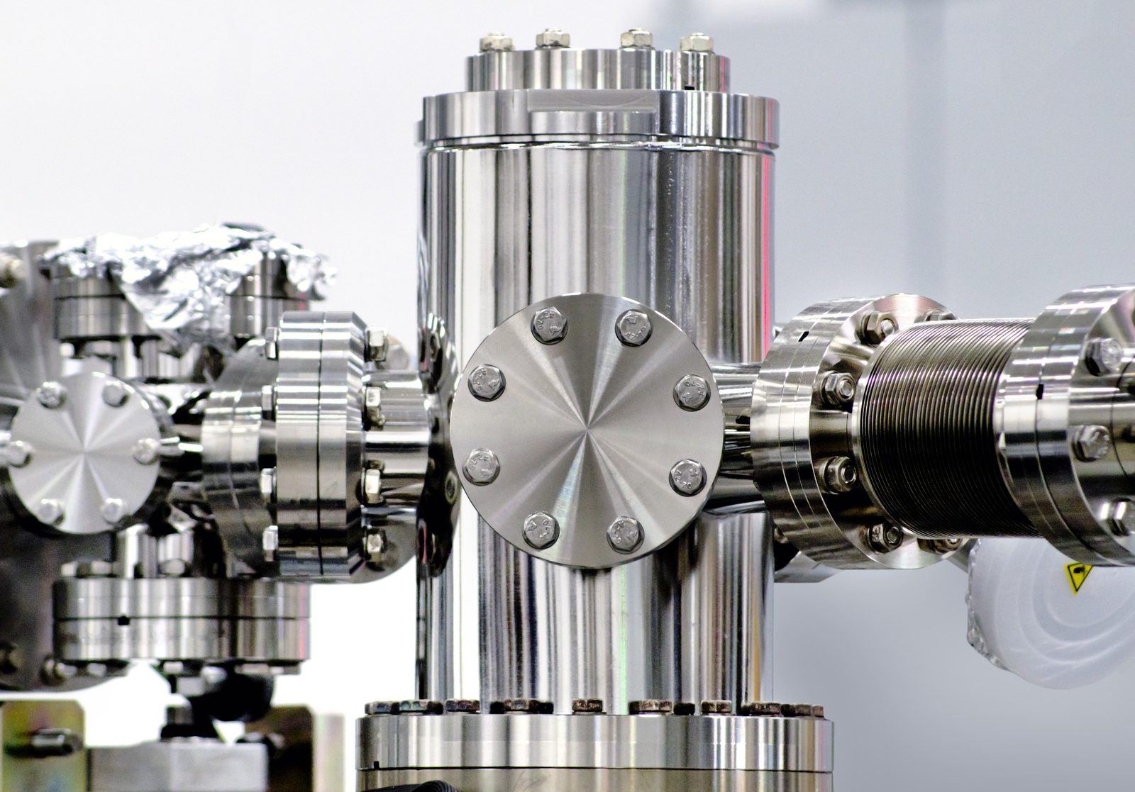 A polished stainless-steel ultra-high-vacuum chamber: a central tee sealed with CF flanges and copper gaskets, a flexible bellows on the right, and aluminium bakeout foil on the upper left.